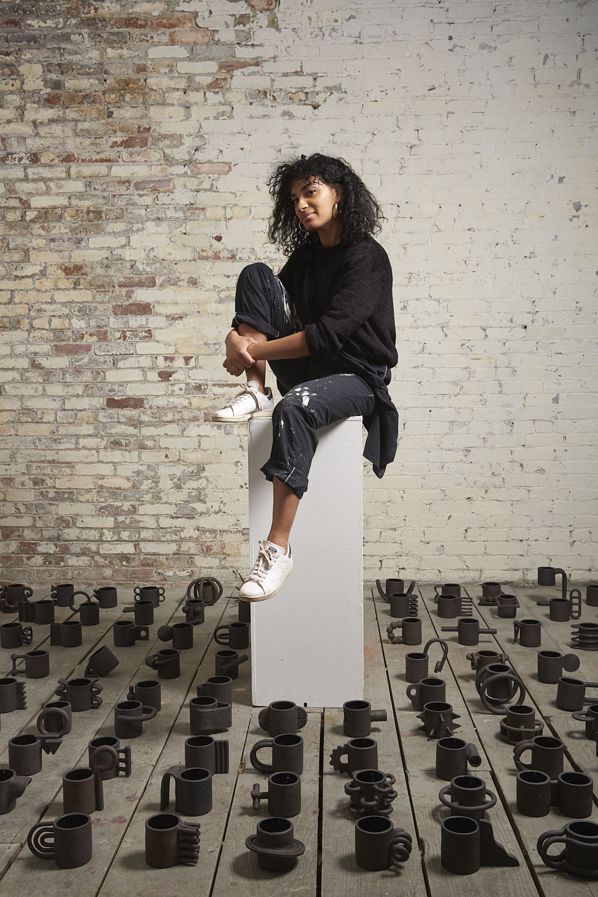 A woman dress in all black with white shoes sitting on a tall white stand. The stand is in the center of a floor filled with black ceramic mugs. 