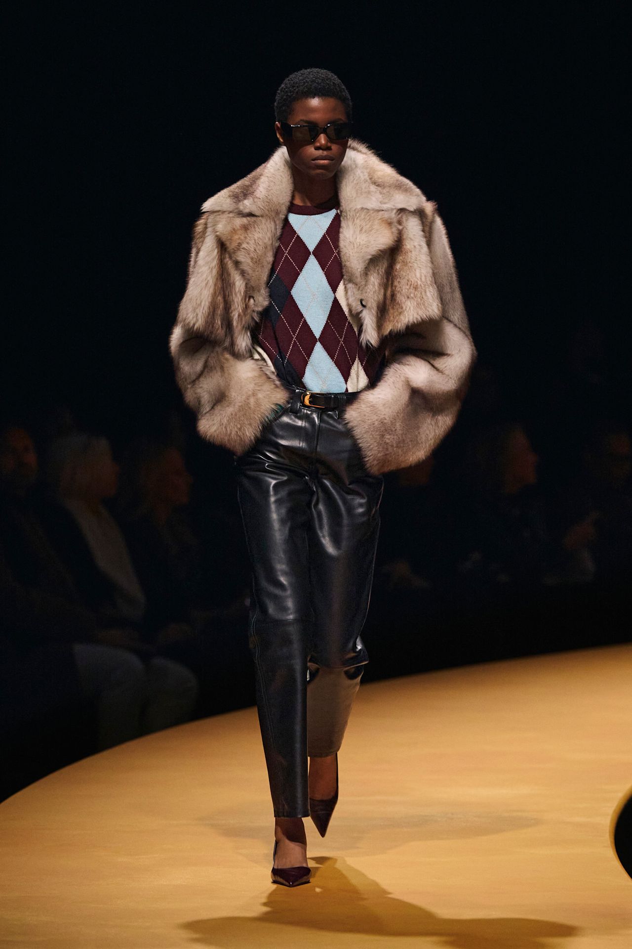 a model at Khaite Fall 2025 wearing a red and blue argyle sweater, gray fur coat, black pants