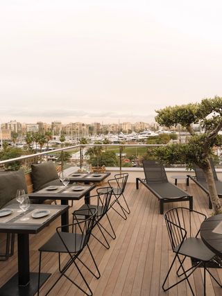 Alternative view of a rooftop area at The Serras featuring wooden decking, a small tree, black chairs, wooden seating with cushions, dark coloured dining tables with tableware and a view of nearby buildings and boats