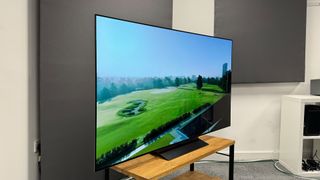 65-inch LG C4 TV photographed at a slight angle on a wooden stand. On the screen is an aerial image of a golf course.