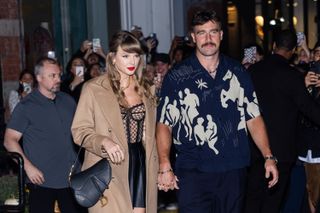 Taylor Swift wearing a tan coat with a sheer black corset holding hands with Travis Kelce in front of a fan of crowds