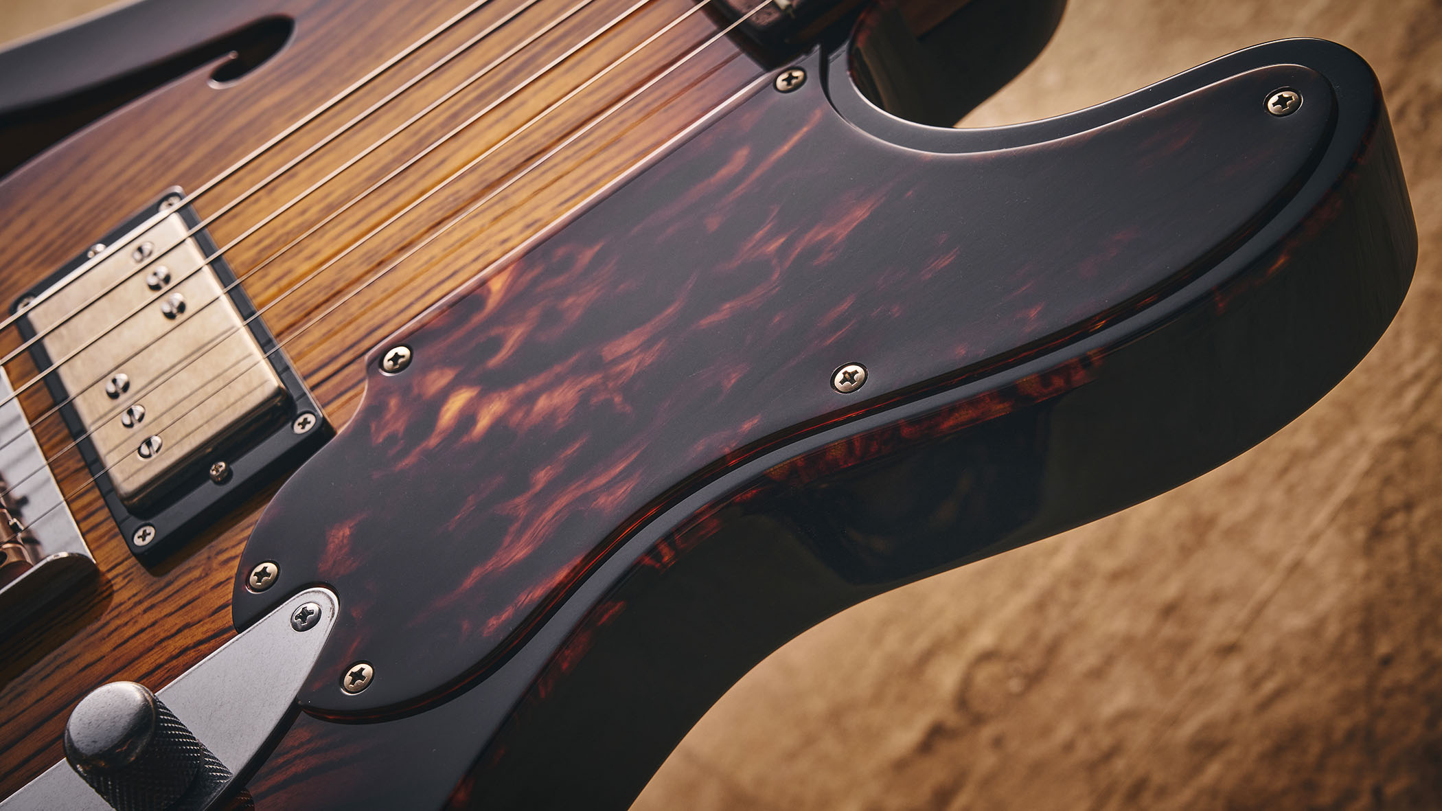 Patrick James Eggle Oz-t Cabronita Thinline; this semi-hollow T-style has a dark sunburst that reveals all the open grain, complementing the tortoiseshell pickguard, and is photographed here against a distressed floor, white and wooden backgrounds.