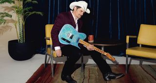 Brad Paisley wears a white cowboy hat, burgundy jacket black jeans and cowboy boots as he sits with his new Fender Lost Telecaster from the Custom Shop.