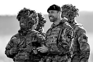 A black and white picture of Prince William wearing army fatigues and using a drone while smiling with soldiers