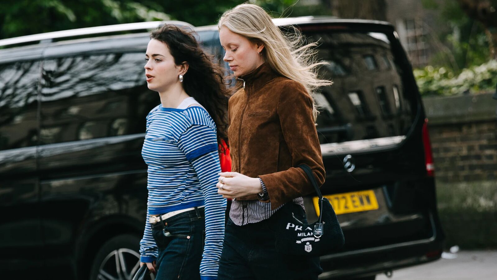 Two woman walk across the street. One is wearing a longsleeve blue striped t-shirt, cuffed jeans, and heels with socks. The other is wearing a brown suede jacket, black jeans, and Adidas sambas. 