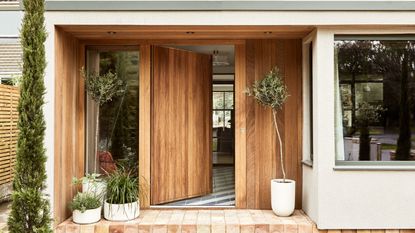 A wooden oak front door opening into a home. Plants in white pots on wither side of the door, red brick porch flooring.