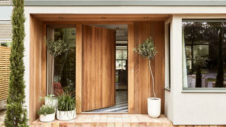 A wooden oak front door opening into a home. Plants in white pots on wither side of the door, red brick porch flooring.