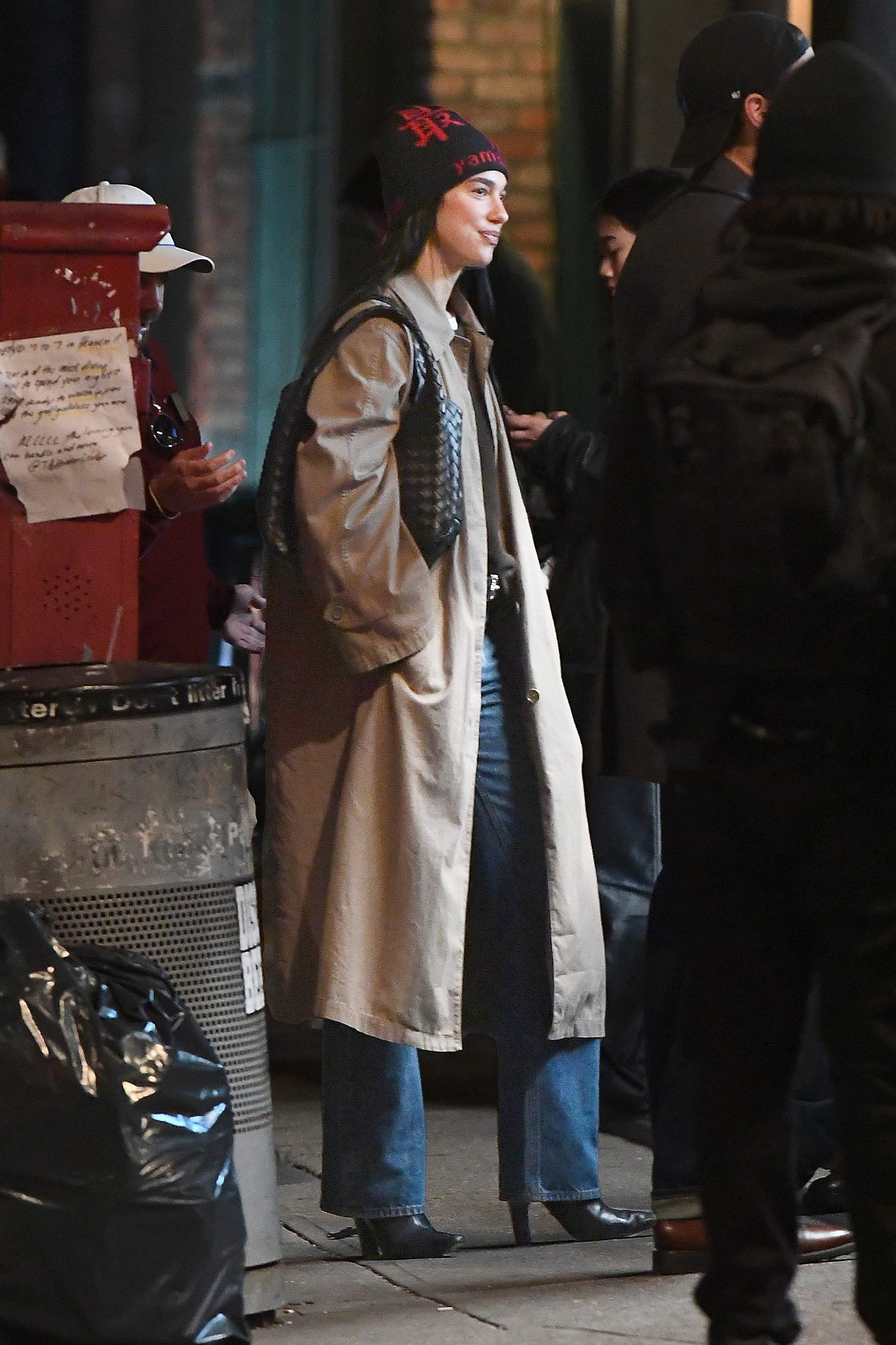 Dua lipa wears a beanie, black bottega veneta bag, beige trench coat, blue jeans, and black heeled boots.