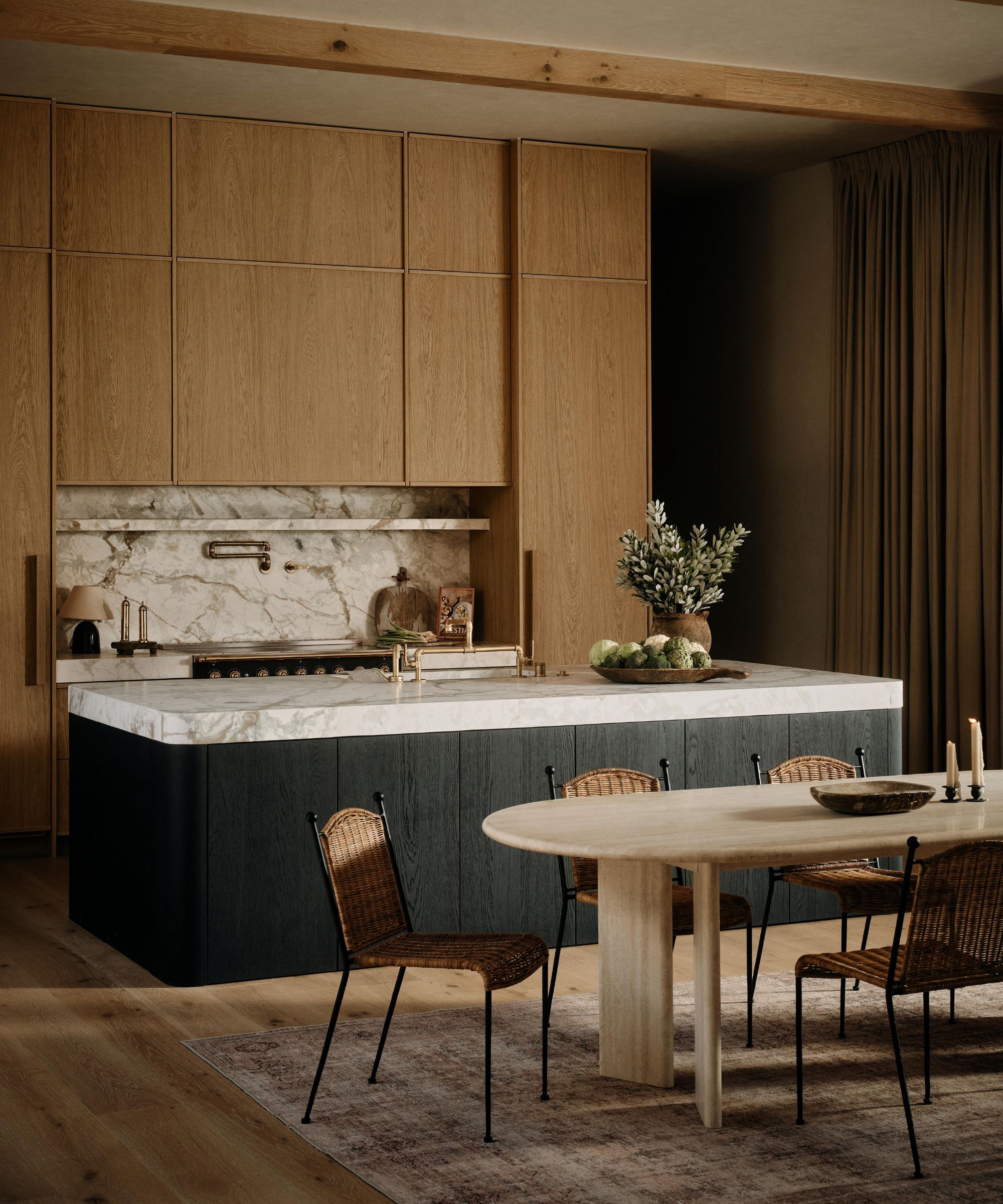 a tall kitchen with oak fronted cabinetry and statement marble stone with a dark island and a travertine dining table and rattan chairs
