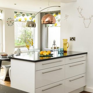 white kitchen with black countertops, white blinds with a yellow, green and grey patten and chrome light fittings