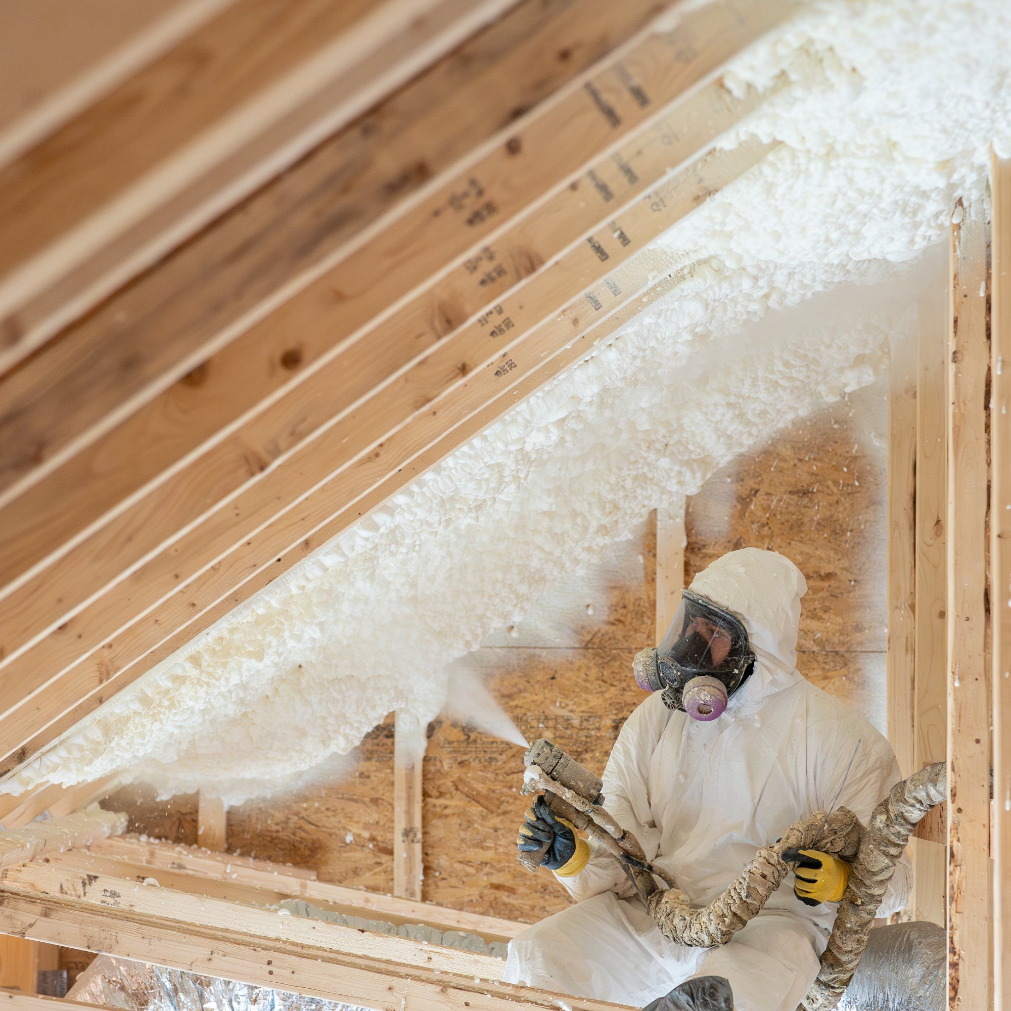 Spray foam insulation being installed in a loft space by a person in white overalls wearing a mask
