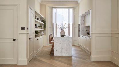 wooden kitchen floor ideas, off white/white kitchen with wooden flooring, marble waterfall style kitchen island, bay window with shutters and voile blind 