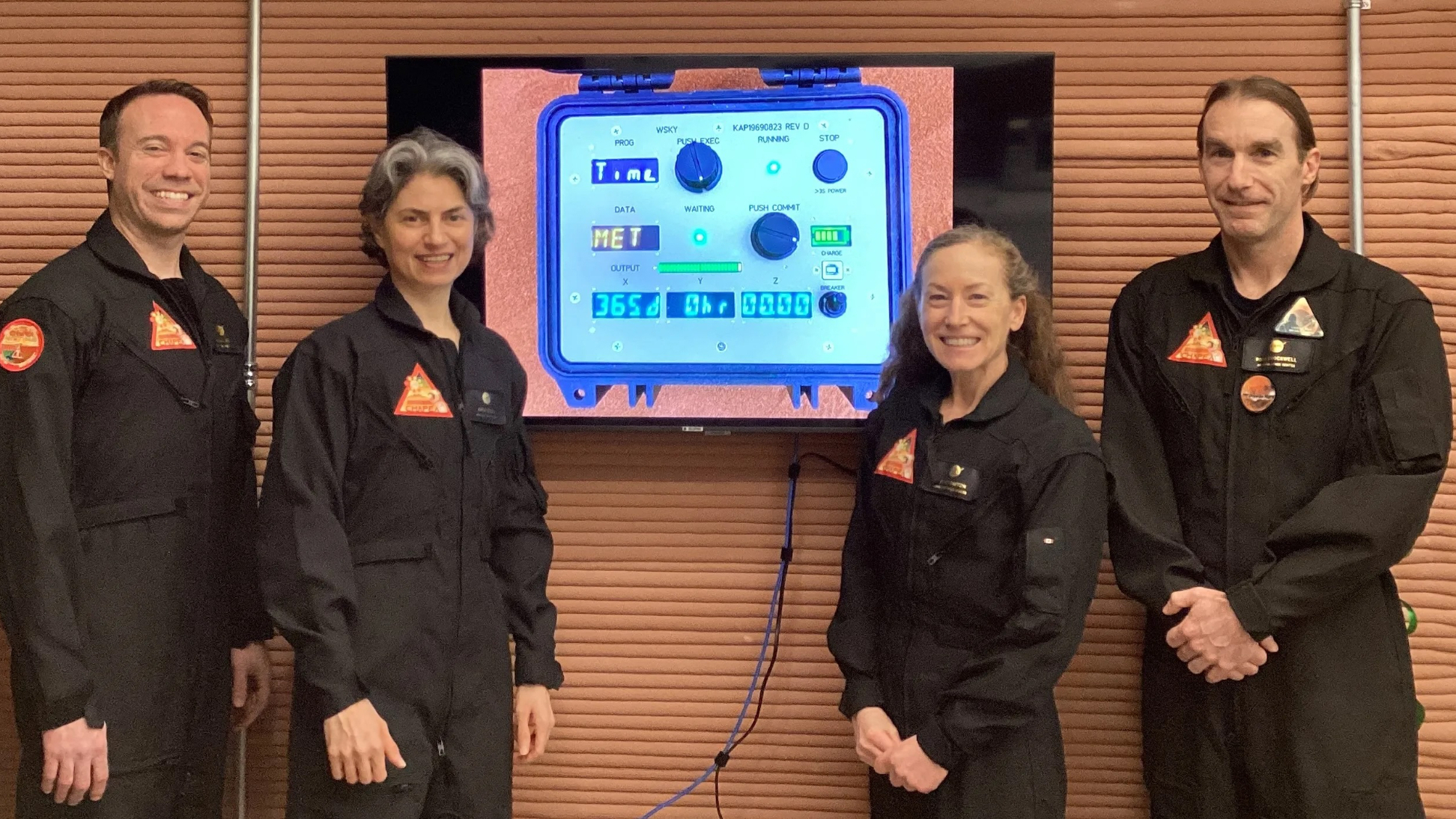 four people in black flight suits stand in front of a tan wall with a tv mounted on it