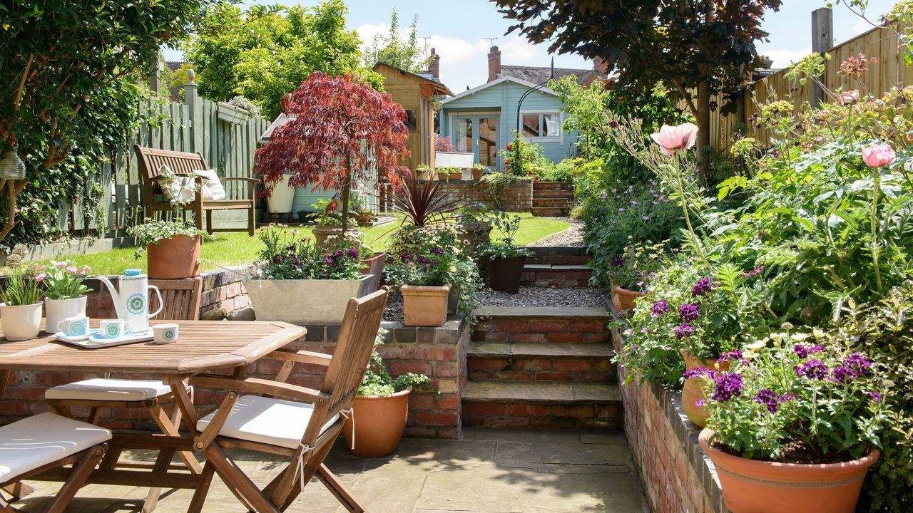 Garden patio with wooden dining table and chairs and lots of potted plants, with steps leading up to grass and a summer house