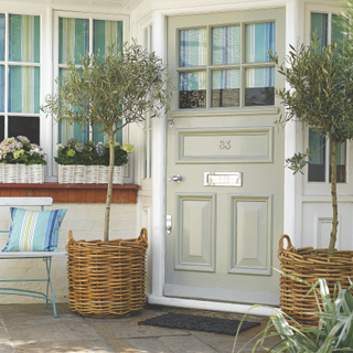 Grey front door with chrome plated door numerals, letter plate, oval door knob, striped blinds, natural wicker baskets, vintage French folding basket, olive trees, window boxes, potted geraniums and hydrangeas