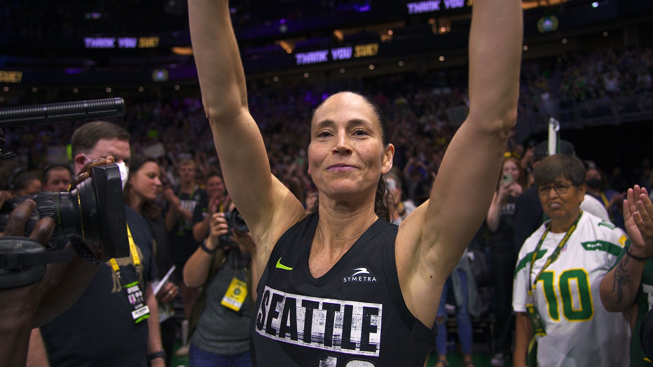 a woman (sue bird) wearing a seattle basketball jersey raises her hands while standing in front of a crowd
