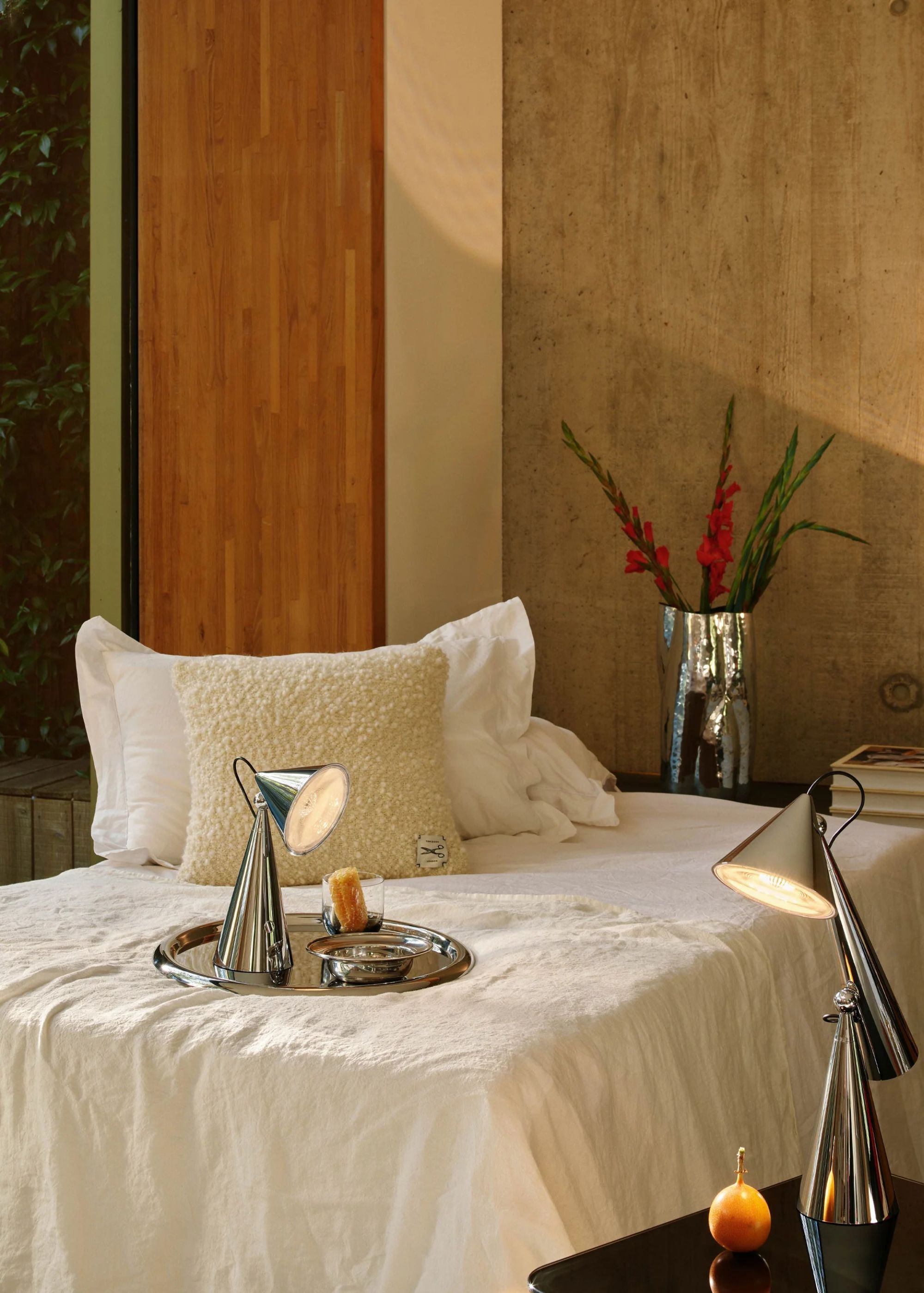 An outdoor lamp sits on a tray on top of a sun bed. There is also a chrome colored vase with plants inside