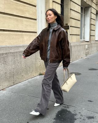 Viki wears a brown leather bomber jacket, gray turtleneck, gray pants, white flats, and an ivory east-west bag.