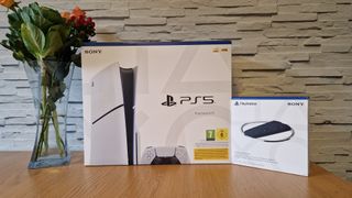 The PS5 Slim and vertical stand in their boxes on a wooden table with a white brick background and next to a vase of flowers