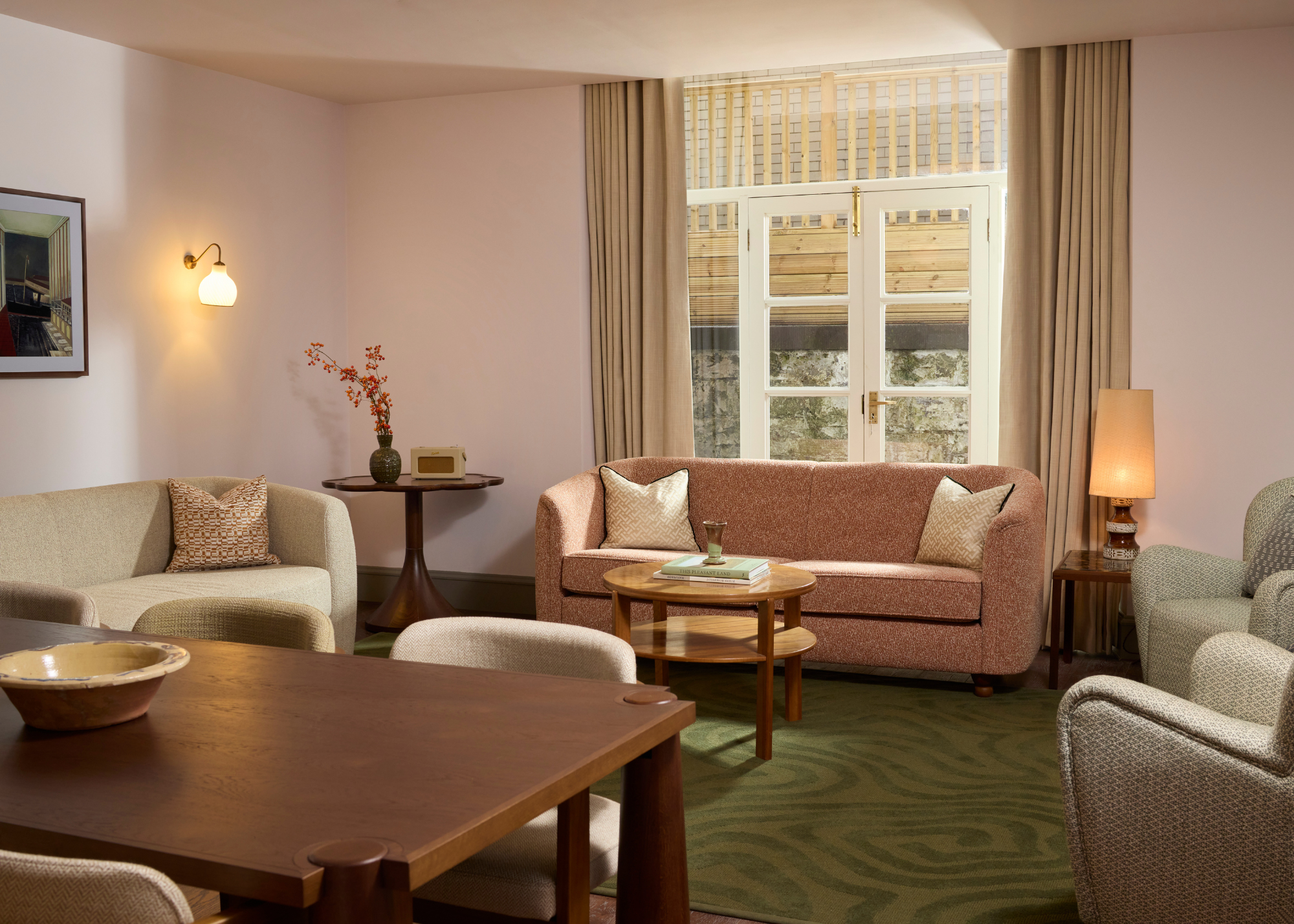 A living room with a green area rug, a wood dining table, a pair of couches in greige and brown, by two gray armchairs, and wood side tables with a vase, a speaker radio, and a table lamp
