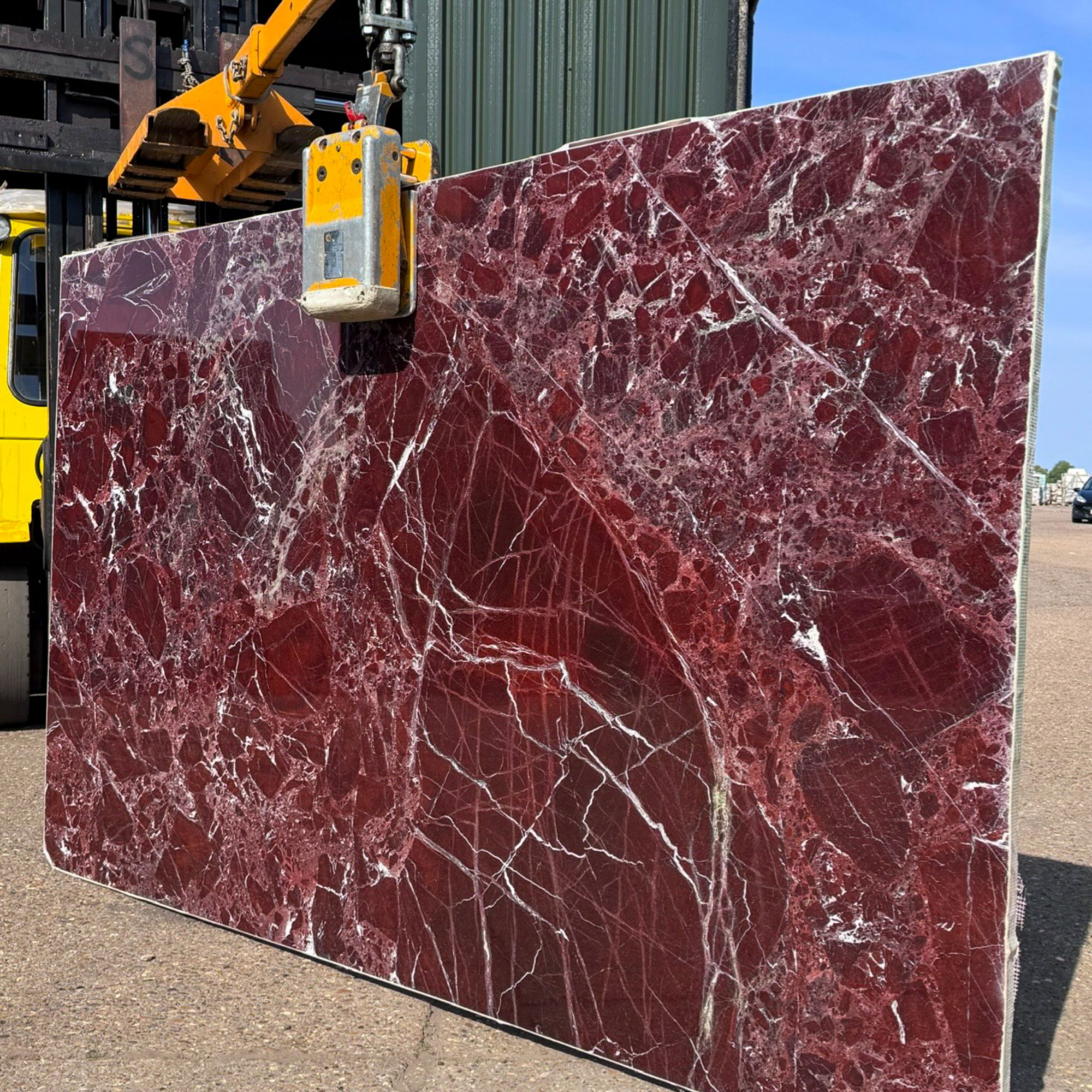 Large slab of deep red marble being held up by machinery in warehouse yard