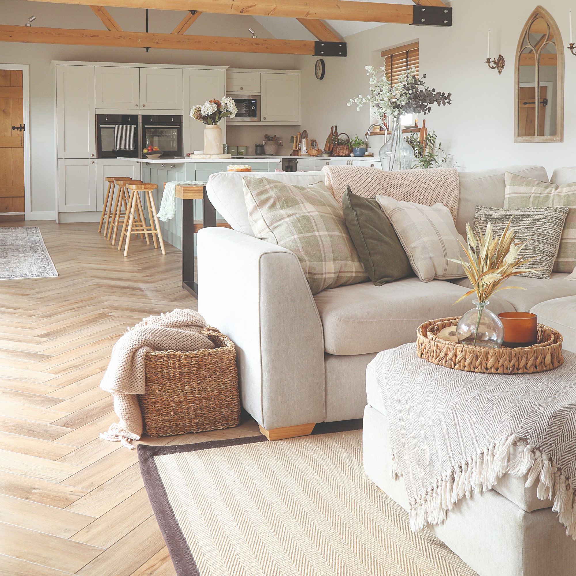 An open-plan living room with a cream corner sofa with a matching ottoman and layered with cushions and a throw