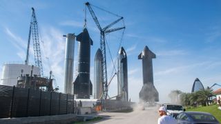 Five towering starship segments stand near cranes against a blue sky, by a dusty road walked on by a guy in a backwards white ball cap and tshirt.