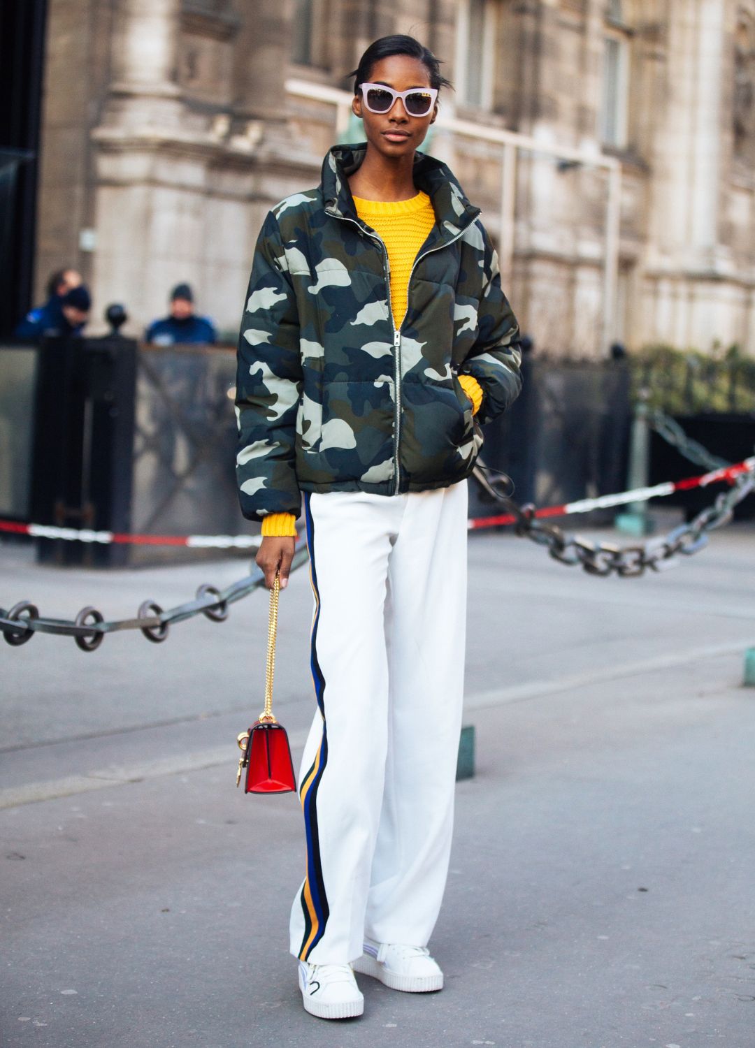 Jamaican model Tami Williams wears a camouflage green jacket over a yellow sweater, white striped Zara trackpants, white sneakers, a red purse, and pink lucite sunglasses on February 28, 2018 in Paris, France