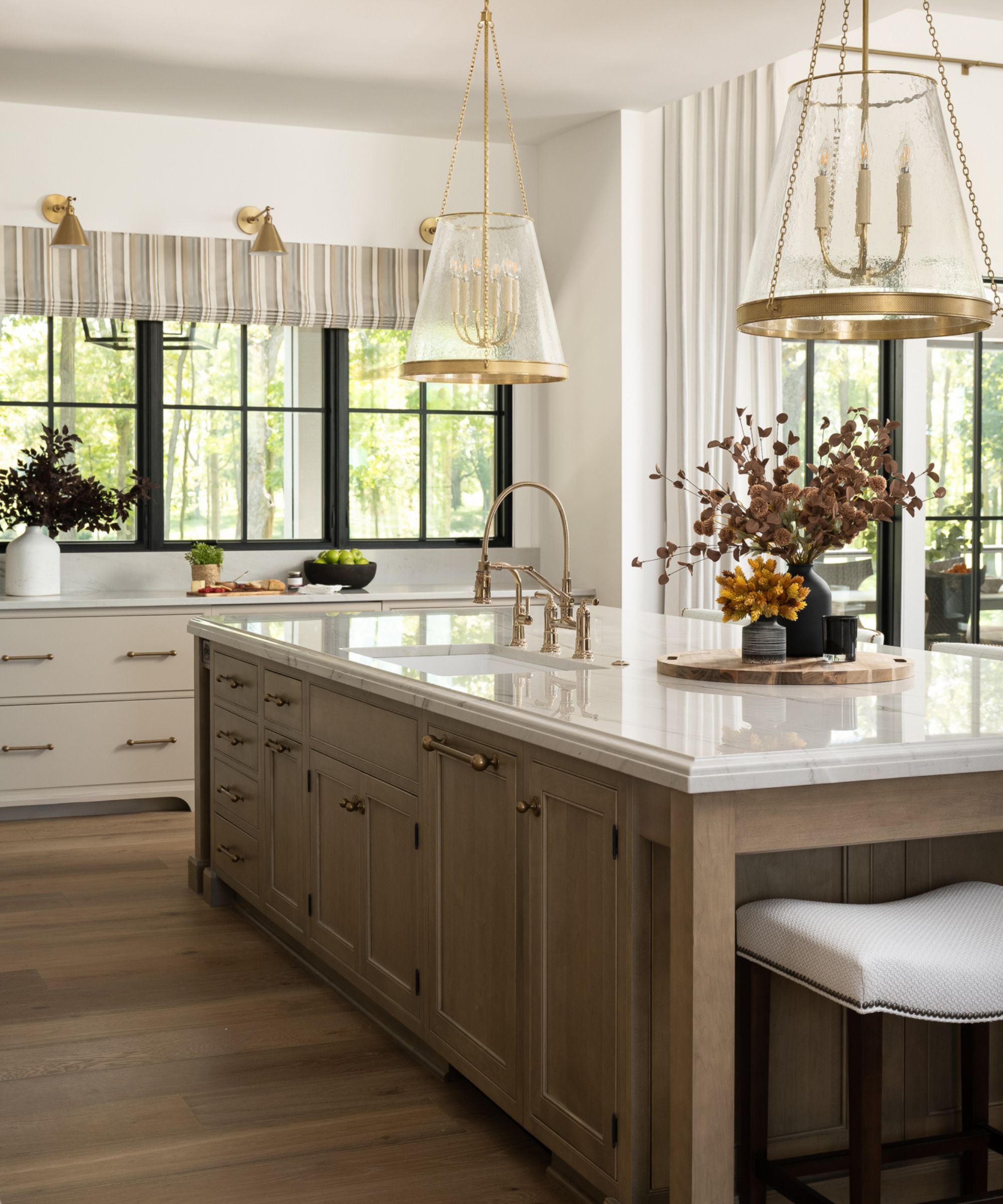 A vertical shot of a large wooden kitchen island with a white marble countertop, gold hardware, and large glass pendant lights set against black-framed windows.