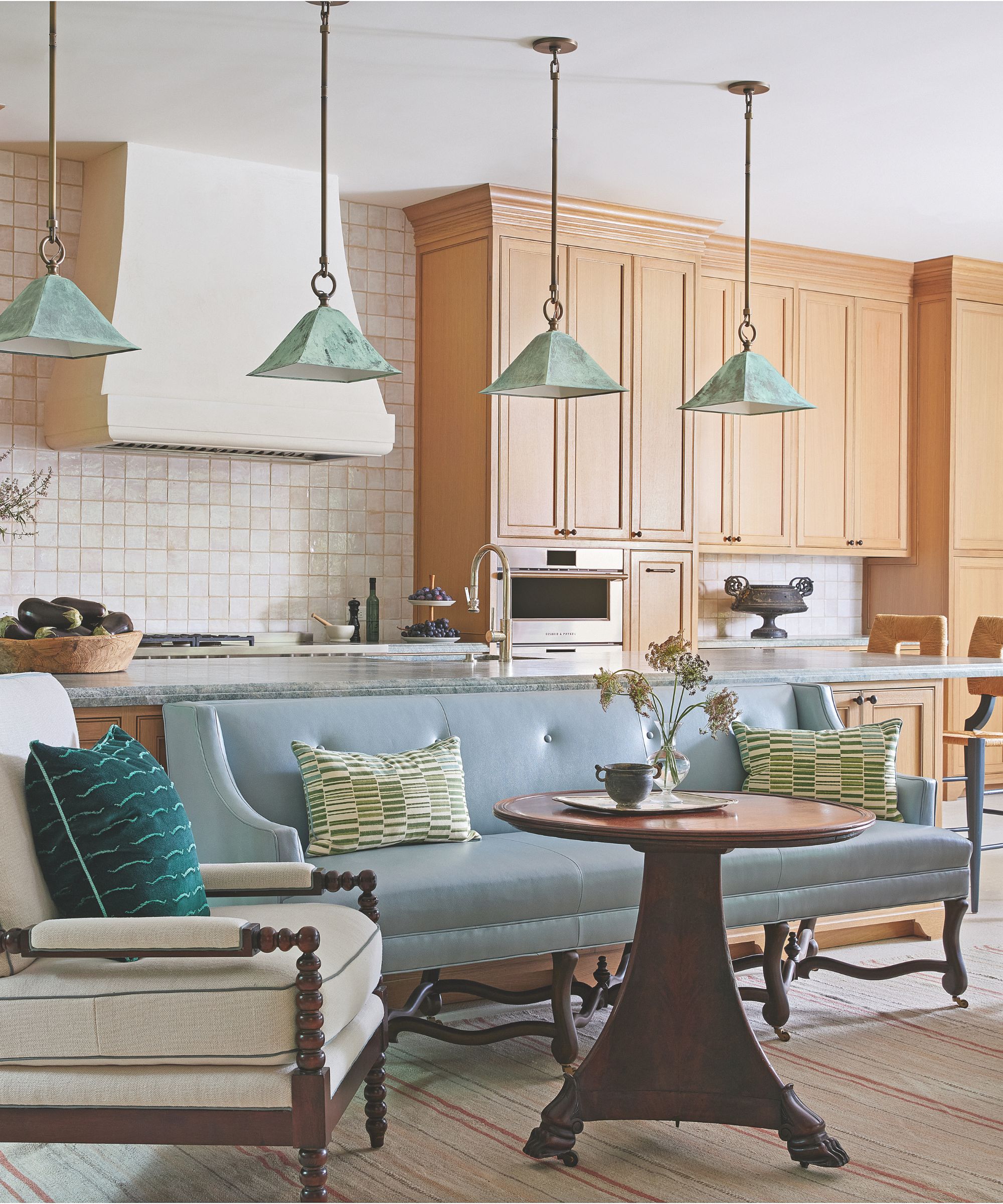 A light-filled kitchen featuring natural wood cabinetry and a marble island, with a light blue leather banquette and an ivory armchair in the foreground under green patina pendant lights