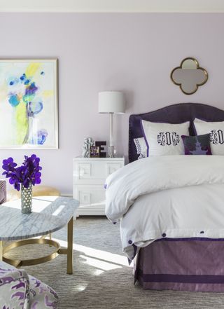 Bedroom with double bed with upholstered headboard and valance and mirror above, nightstand, coffee table and armchair with pale purple wall and neutral carpet