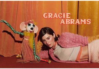 A photo of Gracie Abrams for Who What Wear's fall issue wearing a pink, orange, and yellow look from Miu Miu F/W 24 collection on a stage in a marionette theatre next to a puppet. The artist's name is printed on the photo in pink-and-red capital letters.