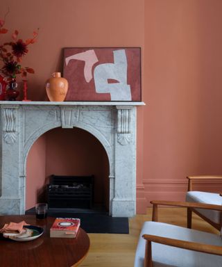 Terracotta living room with color drenched walls and a marble fire surround with artwork positioned on top, an orange vase and a round wooden coffee table