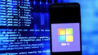 Microsoft Windows 11 logo on a smartphone set against a background of neon blue code on a screen to denote a cyber security theme