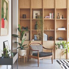 A corner of a living room with built-in shelves displaying books and decorative objects with an accent chair in front of it, surrounded by houseplants