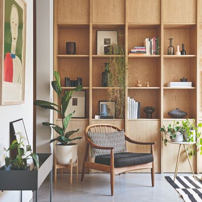 A corner of a living room with built-in shelves displaying books and decorative objects with an accent chair in front of it, surrounded by houseplants