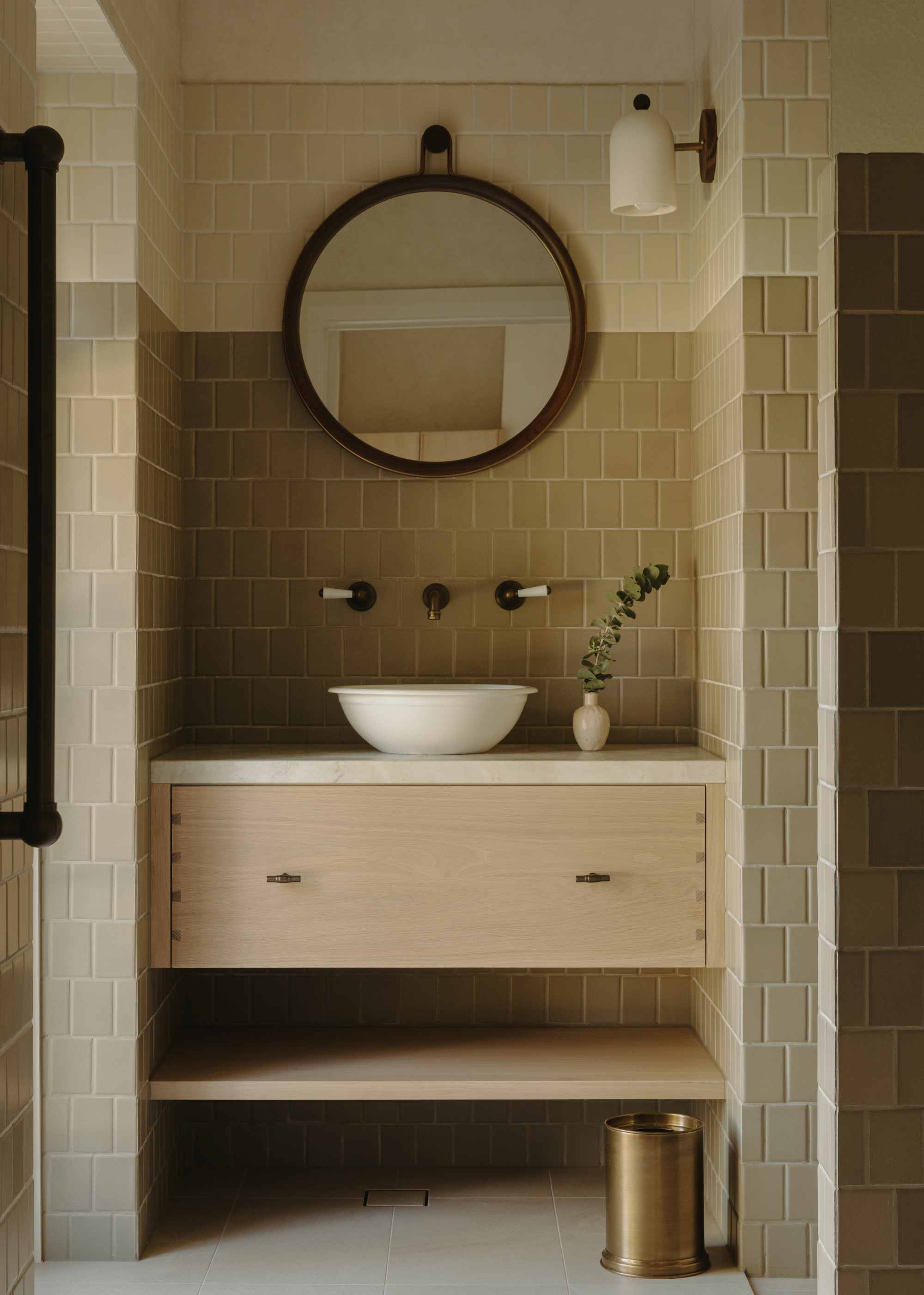 A bathroom with greige and cream tiles, a round hanging mirror, a wall sconce, a white sink, a wooden drawer, a brass trash can and a vase with a stem