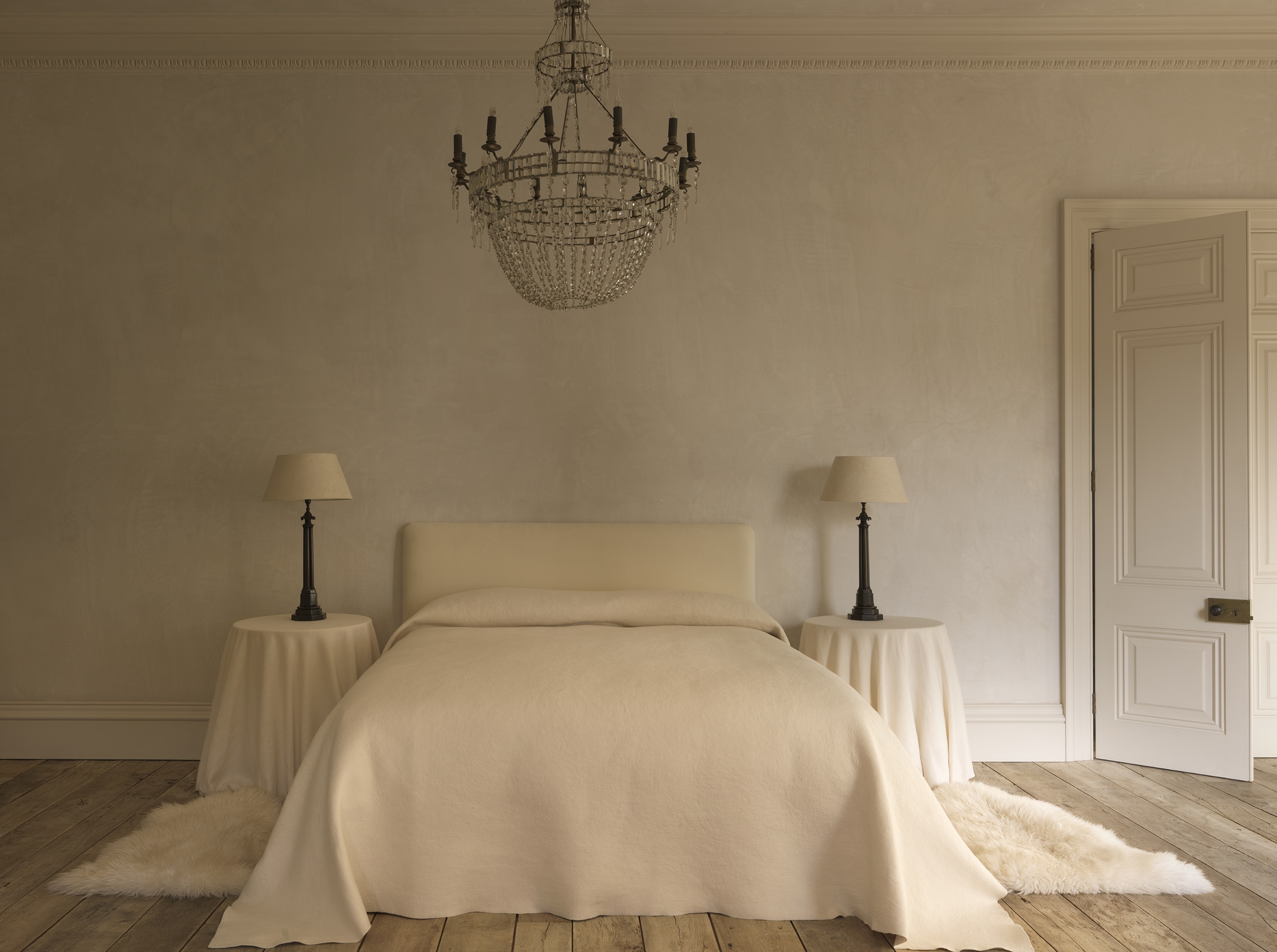 a white bedroom with side tables covered in sheets with a decorative chandelier and limewash walls