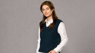 A posed shot of Zahra Ahmadi as DS Esther Williams in Beyond Paradise, visible from the waist upwards. She is standing in front of a grey background and wearing a white shirt with a black sleeveless jumper over the top. She is half-smiling and has her left hand tucked into her trouser pocket while her right arm hangs by her side.