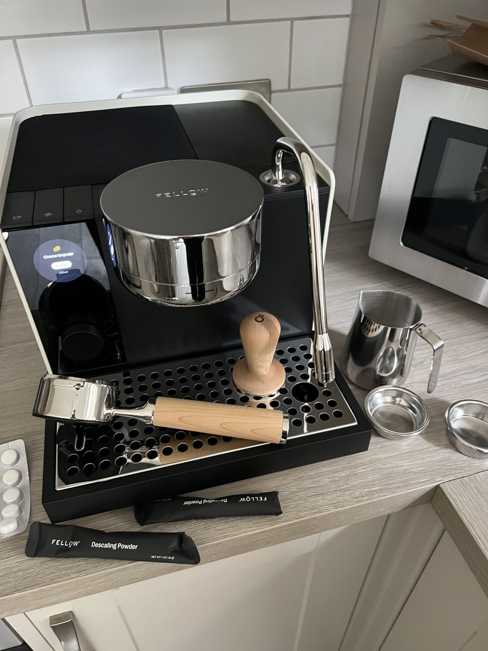 Image of the Fellow Series 1 Espresso Machine on a light wood countertop in a white kitchen. All of the coffee machine accessories it comes with are beside it.