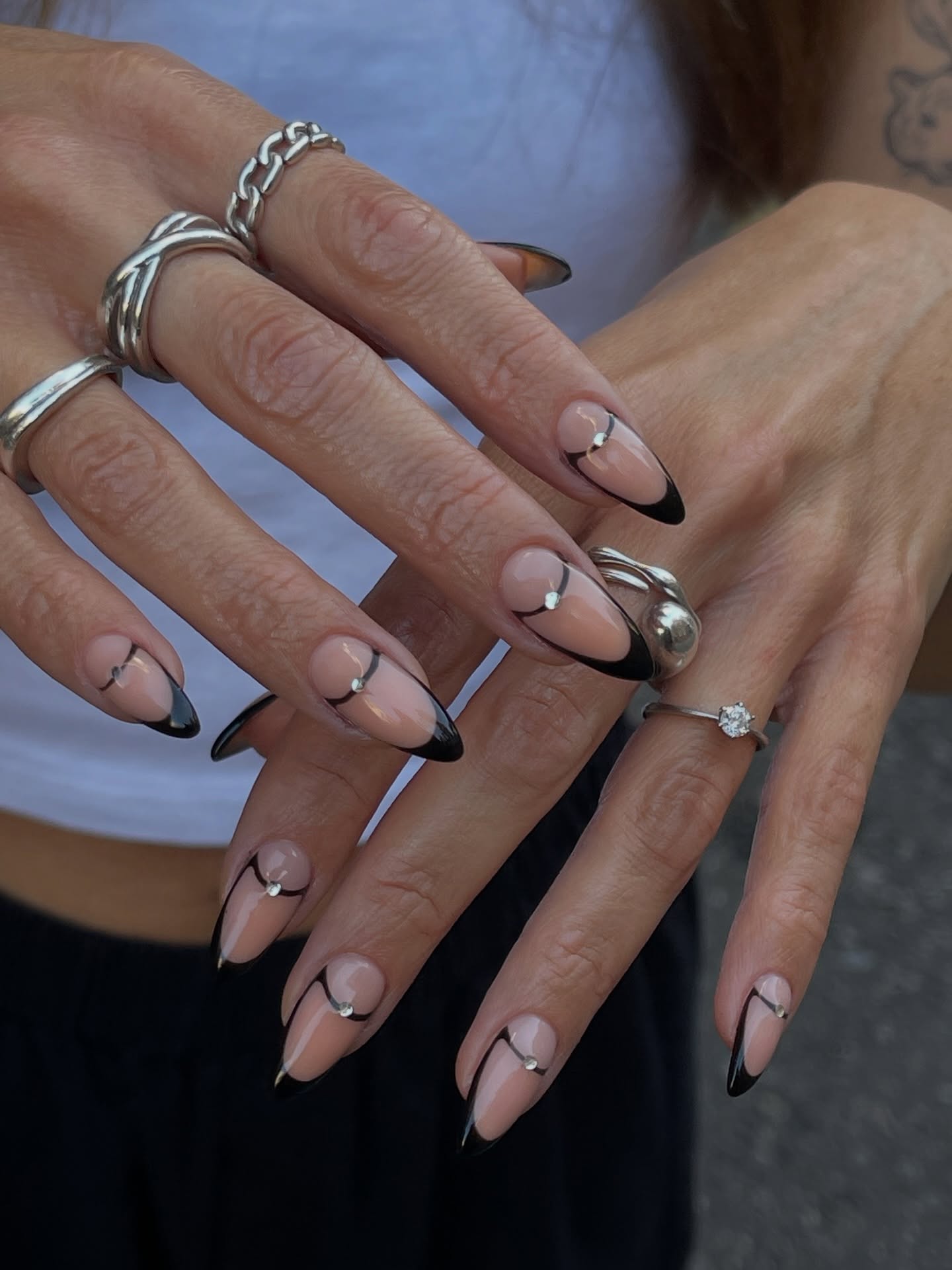 A close-up of long almond-shaped nails with nude polish, black French tips, curved black lines, and small silver accents, paired with silver rings