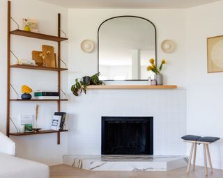 A white living room with white tiled fireplace, arched mirror and floating wooden bookshelves