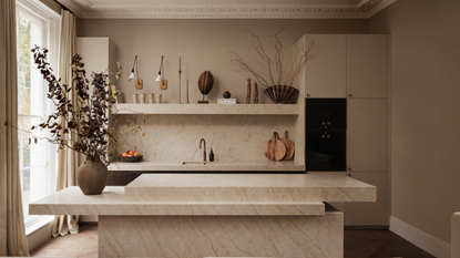 A kitchen with floor-to-ceiling linen curtains, a tiered marble island and a fridge in the corner