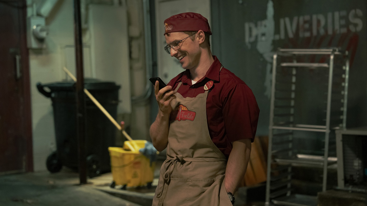Adrian Chase smiling as he speaks on the phone during a lunch break in Peacemaker season 2
