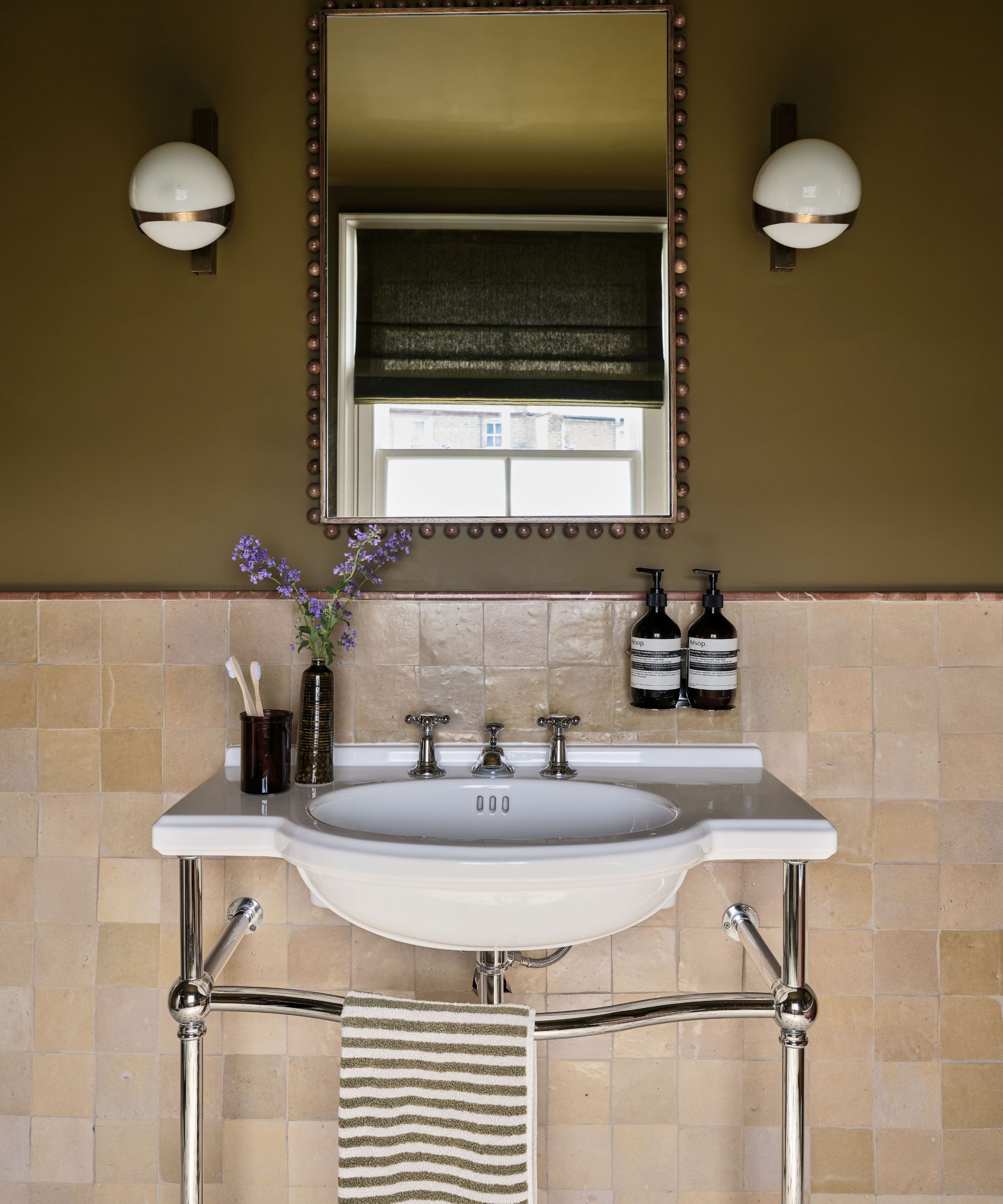 Bathroom with olive green painted wall, beige zellige tiles, freestanding sink with metal frame, a brass rectangular mirror, and round glass sconces either side