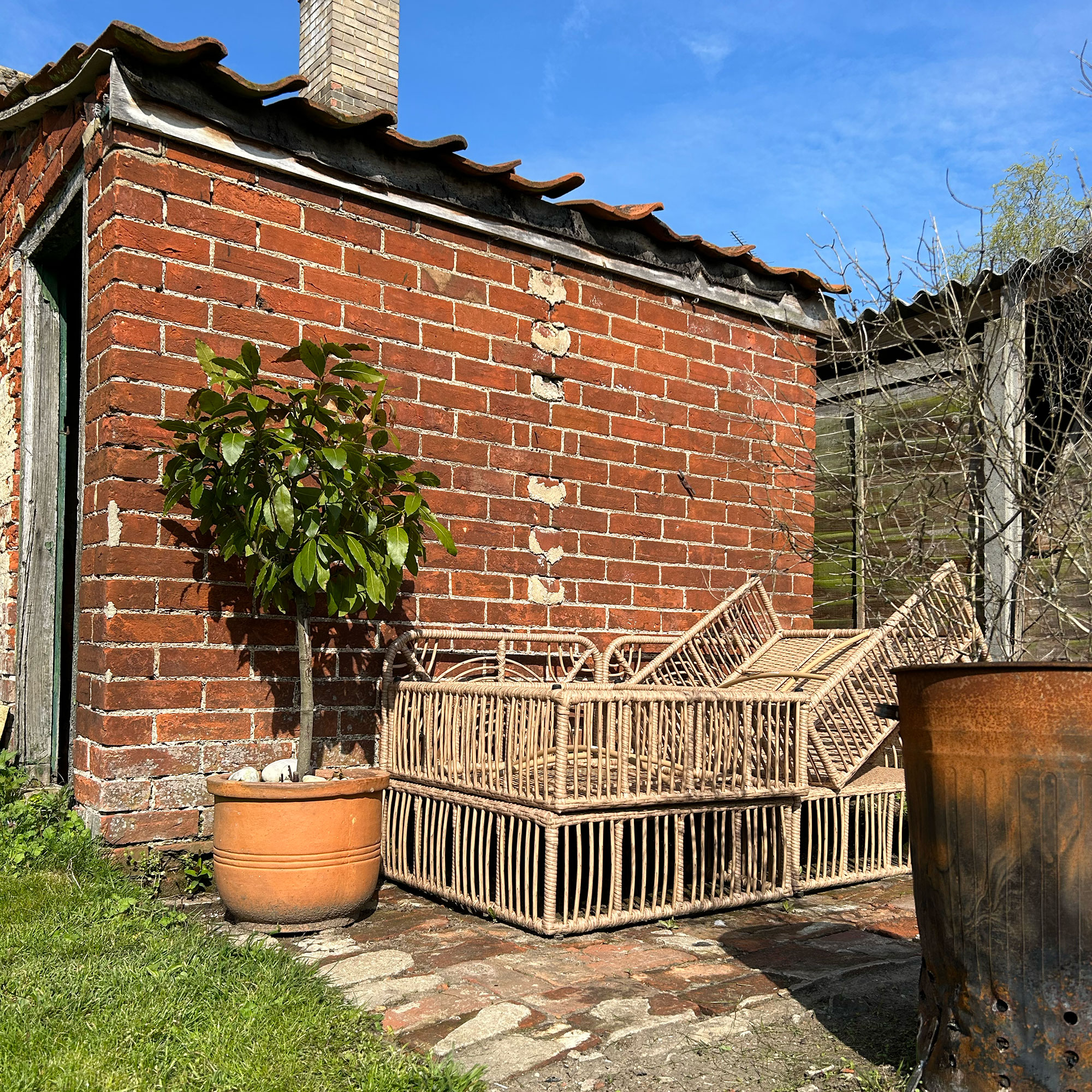 Brick outhouse in garden with patio area beside, holding potted small tree and frames of rattan furniture