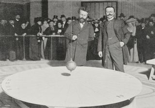 Black and white photo of two men standing in front of a pendulum. A crowd stands behind them