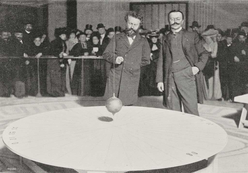 Black and white photo of two men standing in front of a pendulum. A crowd stands behind them