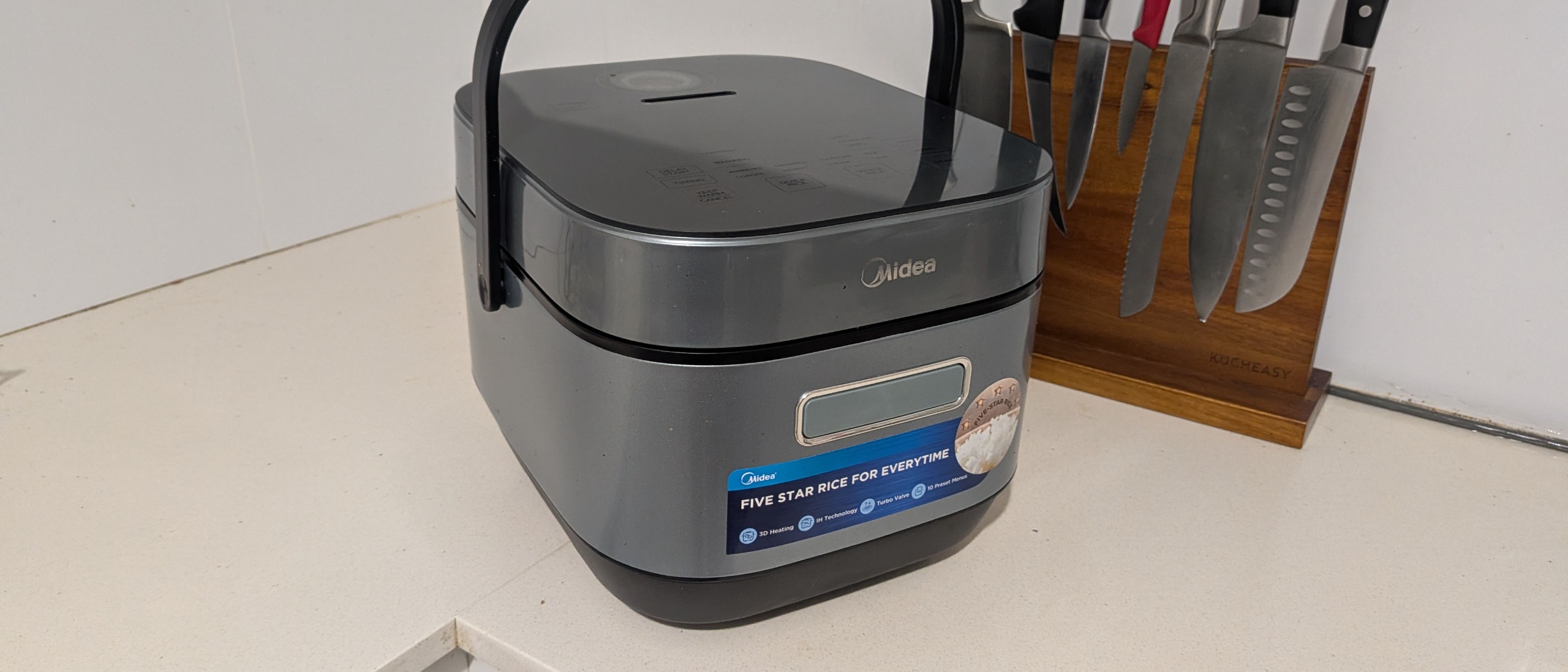 A Midea Induction Rice Cooker on a countertop with some knives on a magnetic knife block