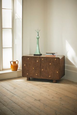 A whimsical wooden cabinet sitting next to an orange amphora vase features a mid-century modern-inspired silhouette brought to life by golden star reliefs. On top of it lie a green sculpture and some coffee table books.